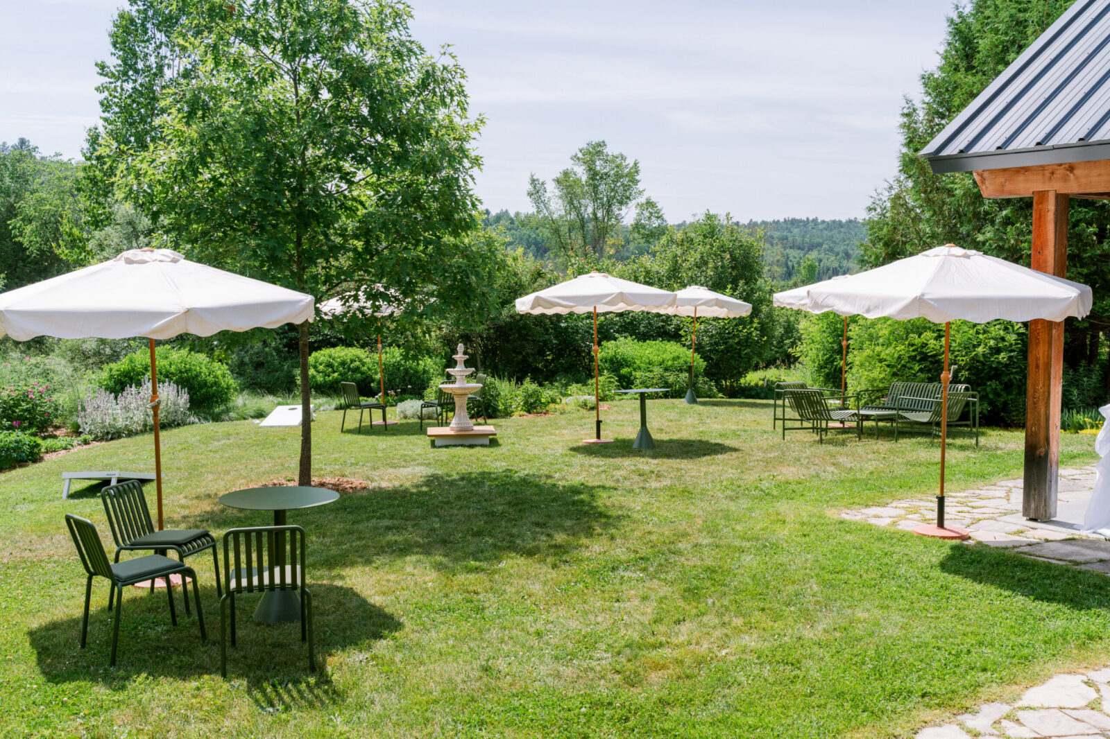 Outdoor lounge area at Le Peaches & Cream with white umbrellas, green bistro chairs, and a stone fountain set on a lush lawn surrounded by trees.