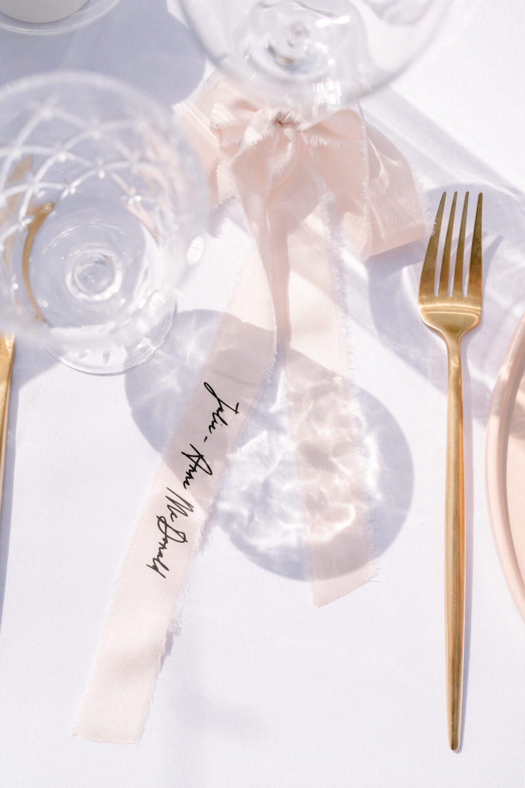 Elegant place setting with a hand-lettered ribbon place card, blush bow, gold flatware, and crystal glassware at Le Peaches & Cream.
