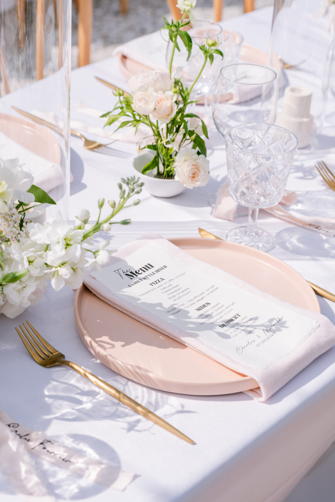 Blush place setting with gold flatware, a printed menu, and delicate white floral sprigs on a soft pink charger at Le Peaches & Cream.