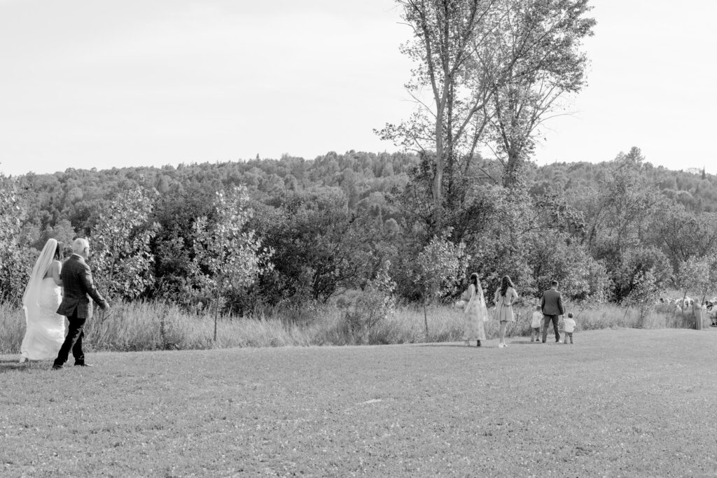 Black-and-white photo of a bride and groom walking hand in hand across an open field at Le Peaches & Cream, with wedding party members and trees in the background.