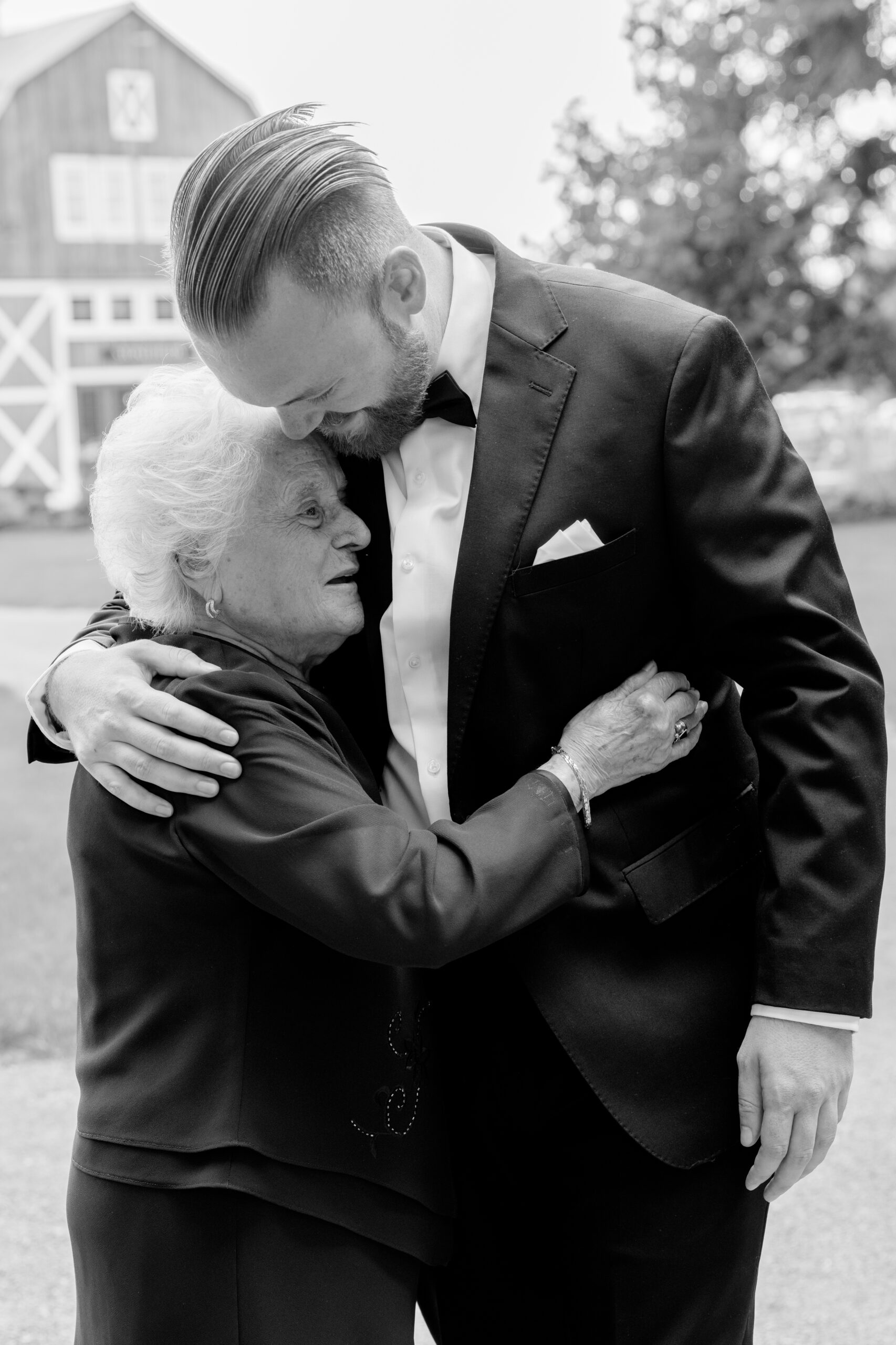 A heartfelt black-and-white image of a man in a suit gently hugging an older woman outdoors. The scene conveys warmth and love, with a barn in the background.