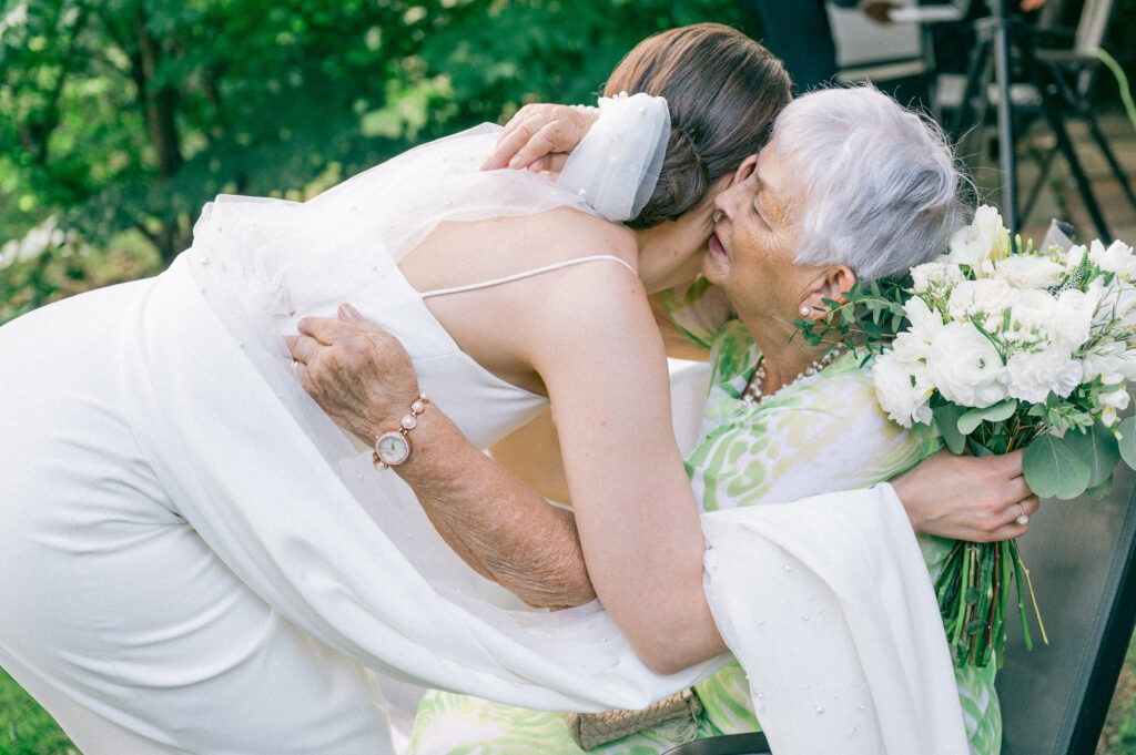 A bride in a white dress embraces an elderly woman holding a white bouquet. The scene is warm and emotional, set against a lush green outdoor background.