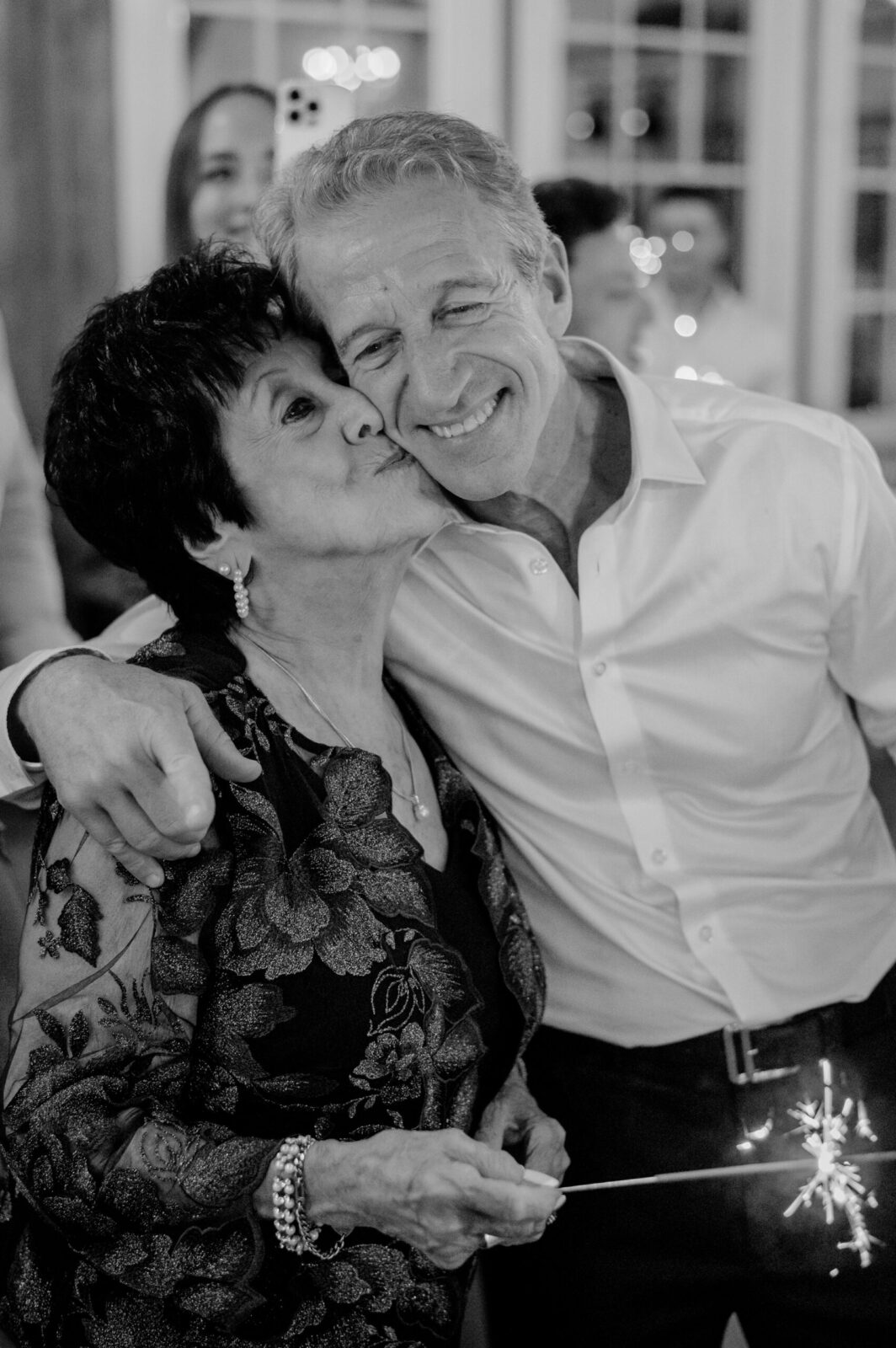 An older woman affectionately kisses a smiling man on the cheek at an indoor celebration. Both hold sparklers, creating a joyful and warm atmosphere.