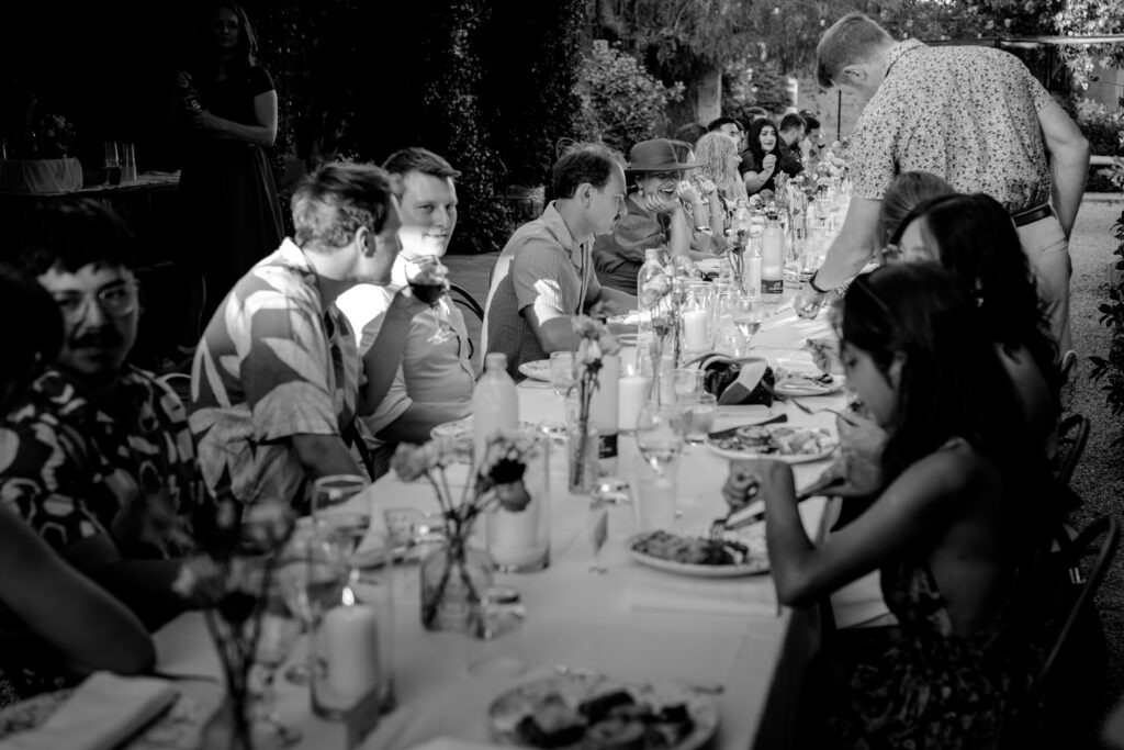 Wedding guests gather around a long candlelit dinner table, sharing conversation during an outdoor evening reception.