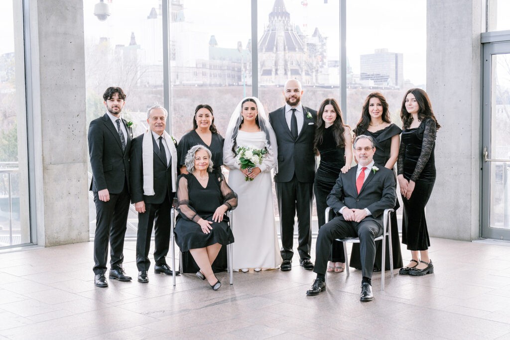 A wedding party family photo with ten people, centered around the bride in a white gown holding a bouquet. Others in black attire are smiling, with a cityscape visible through large windows. Mood: joyful and formal.
