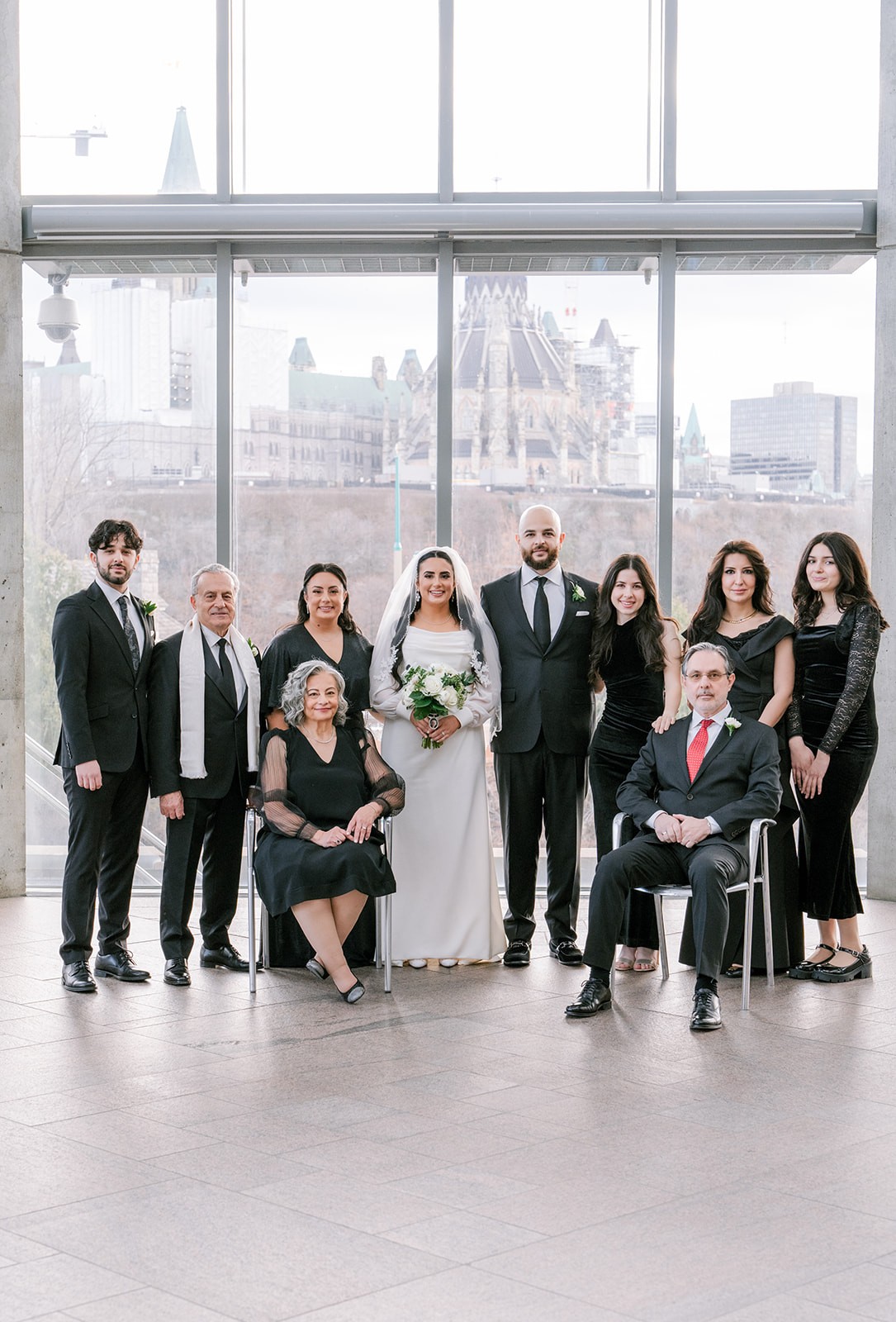 A happy wedding group photo features a bride and groom surrounded by family, all in formal attire. A cityscape is visible through large windows behind them.