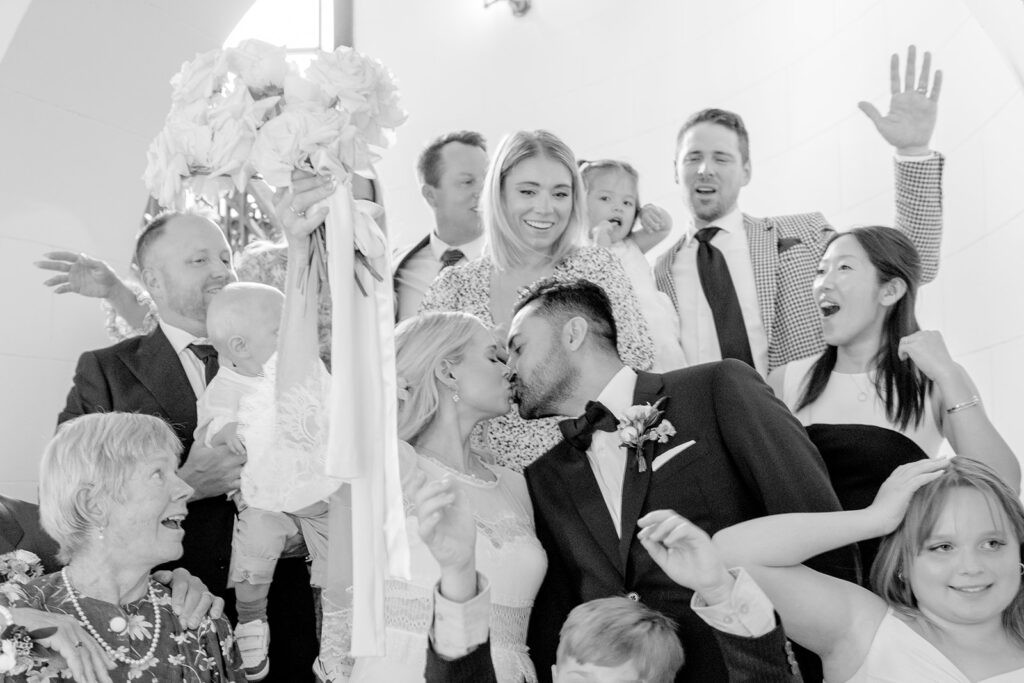 Joyful black-and-white wedding scene with a kissing couple surrounded by smiling family and friends, capturing celebration and love.