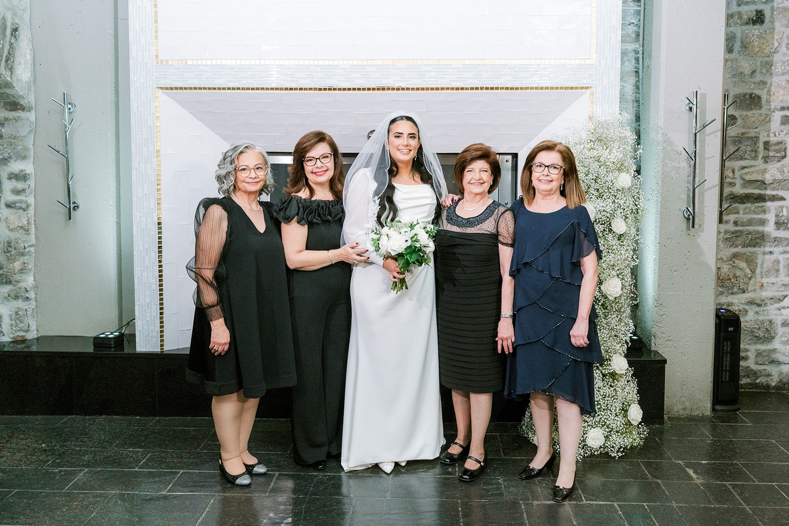 Bride in white dress and veil holds a bouquet, surrounded by four women in elegant black and navy dresses, smiling in a formal setting.