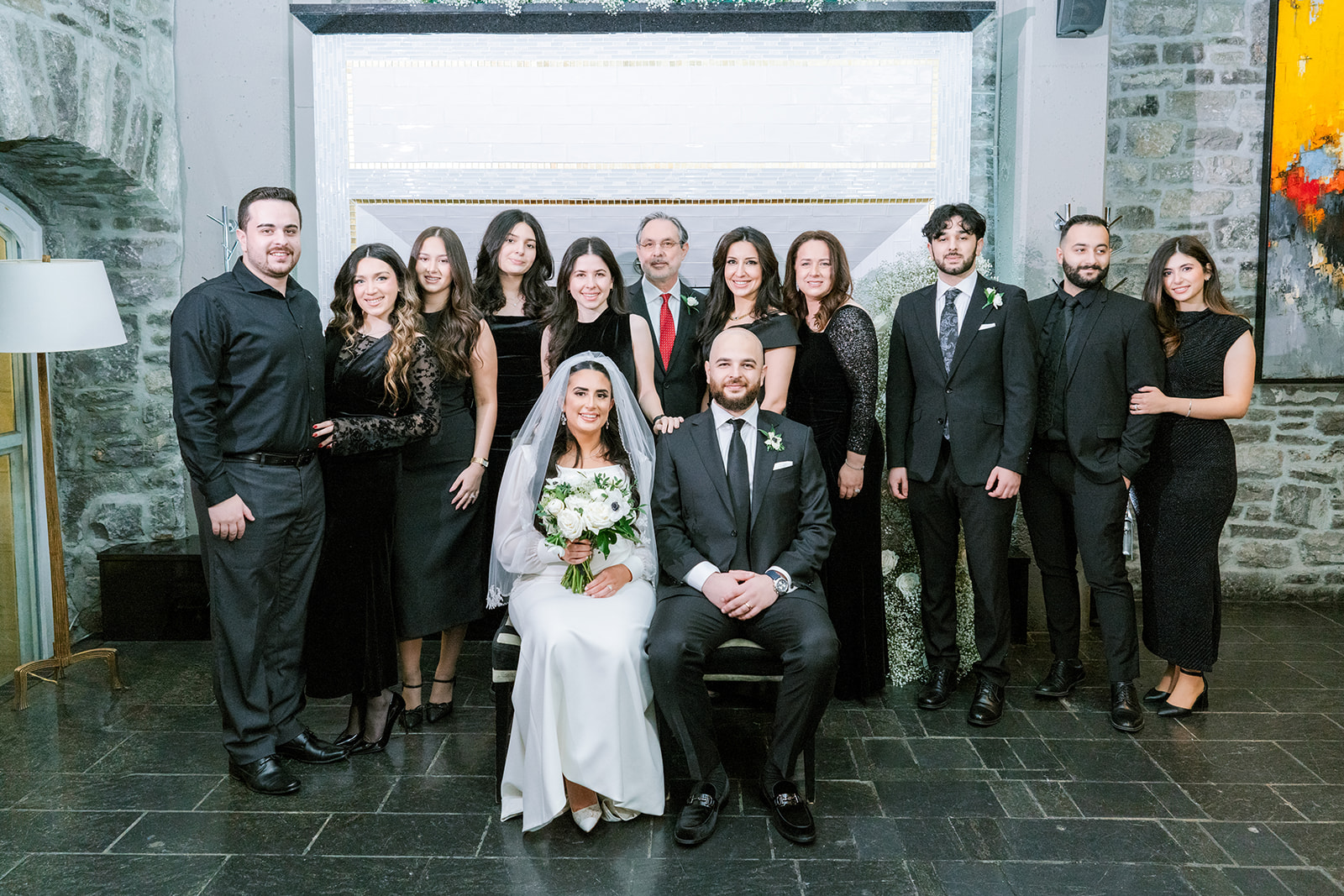 A bride and groom sit at the center of a family group, all dressed in formal wear. The setting is an elegant, stone-walled room, conveying a joyful and celebratory atmosphere.