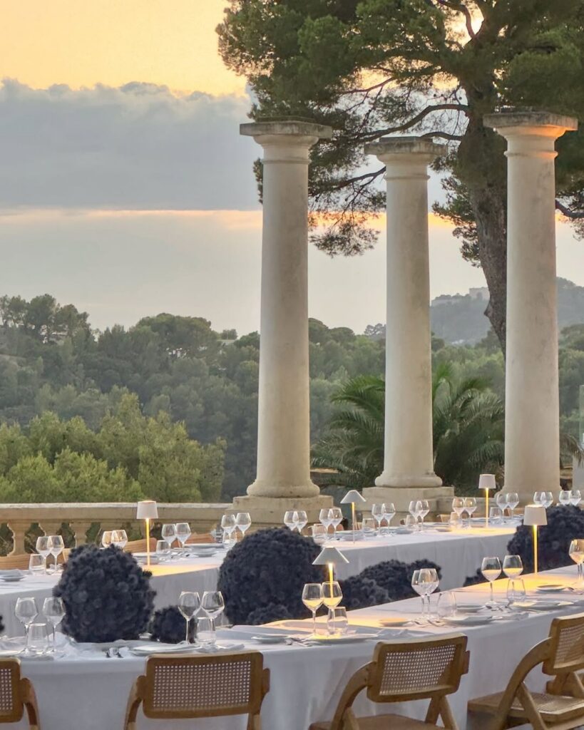 Elegant outdoor dining setup with white tablecloths, blue floral centerpieces, and modern lamps. Columns and lush greenery in the background enhance a serene sunset ambiance.