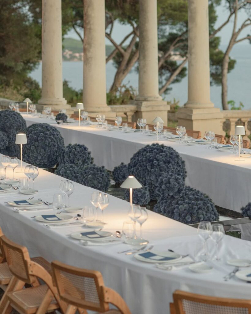 Elegant outdoor dining scene with long, white tablecloth-covered tables, blue floral arrangements, and soft lighting by small lamps, set near classical columns overlooking the sea planned by All Things Beautiful.