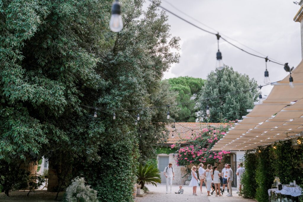 A group of people gather in a lush garden adorned with hanging string lights. A canopy with blooms covers part of the courtyard, adding a festive, relaxed ambiance.
