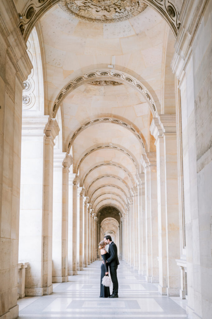 A couple embraces under ornate, arched ceilings in a grand, light-filled corridor, conveying romance and elegance in a historic architectural setting.