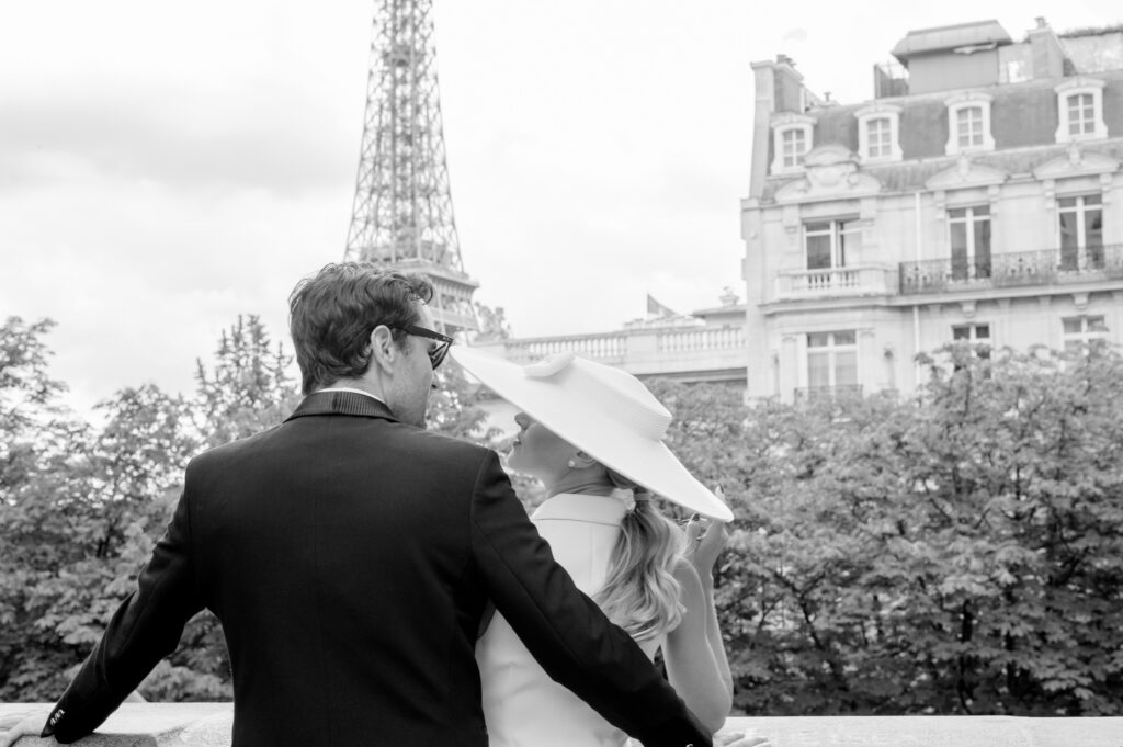 A couple in elegant attire, facing away, leans on a Parisian balcony with Eiffel Tower in view. Romantic, timeless feel in black and white.