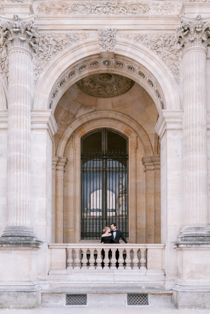 A couple, elegantly dressed, stands embraced on a stone balcony of an ornate, classical building with tall columns and an arch. The scene exudes romance.