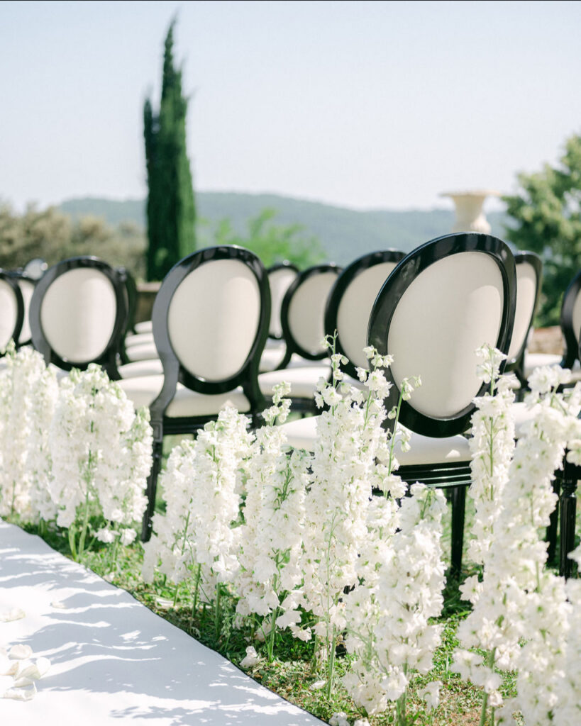 Rows of elegant black and white chairs line an outdoor aisle adorned with white flowers, set against a backdrop of lush greenery and clear sky, planned by Ajna of Oui Mon Coeur.