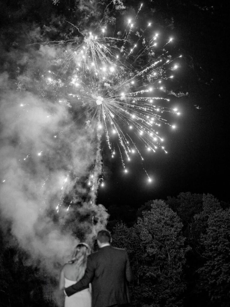 A couple embraces under a dazzling fireworks display in a night sky, surrounded by trees. The scene conveys romance and celebration.