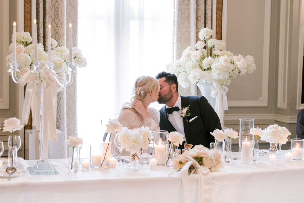 A bride and groom share a tender kiss at their elegantly decorated wedding table, adorned with white flowers and lit candles, evoking a romantic ambiance.
