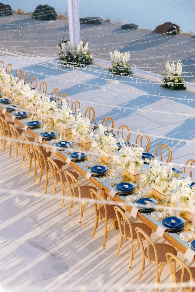 Elegant outdoor wedding table set for a gathering with wooden chairs, blue plates, and white floral centerpieces under twinkling string lights.